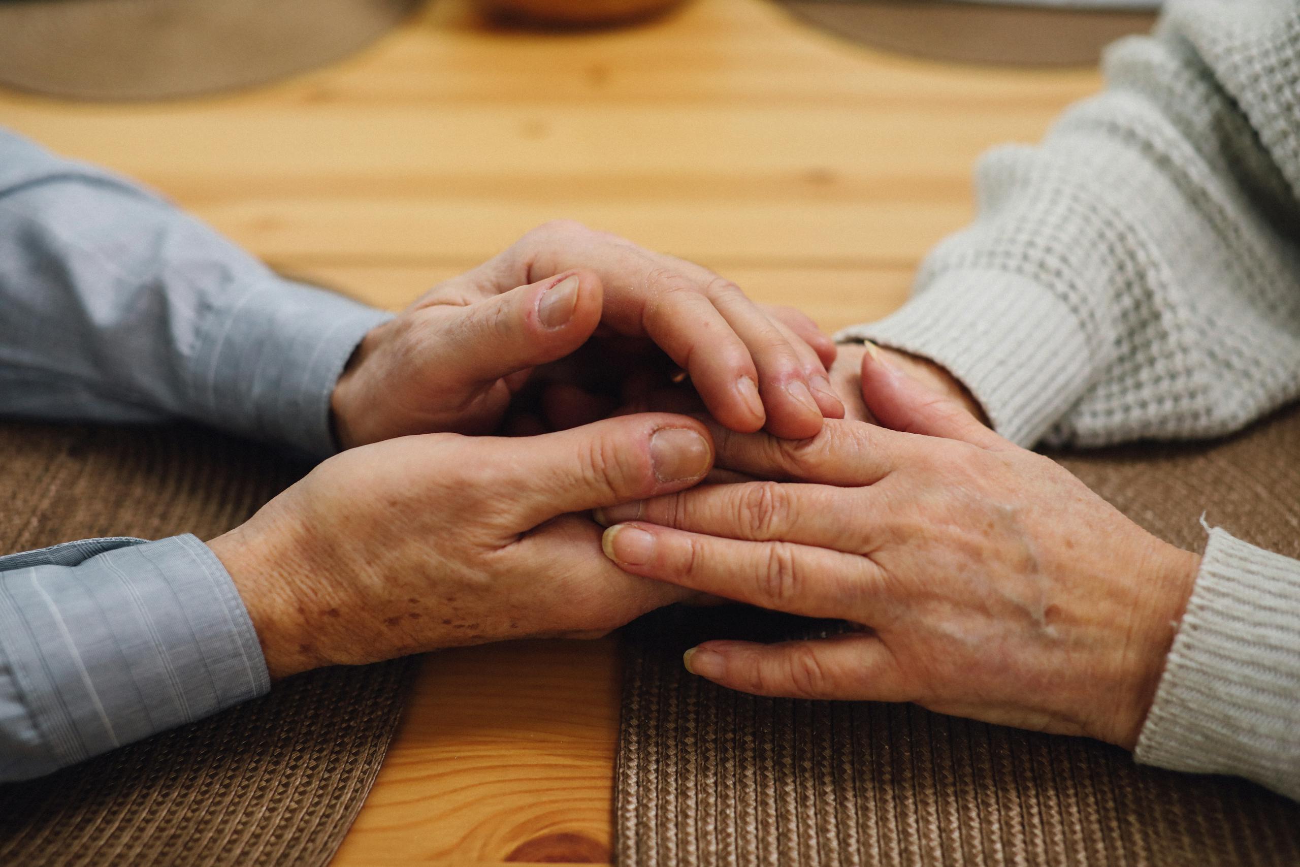 A close-up of an elderly couple holding hands on a wooden table, signifying love and companionship.